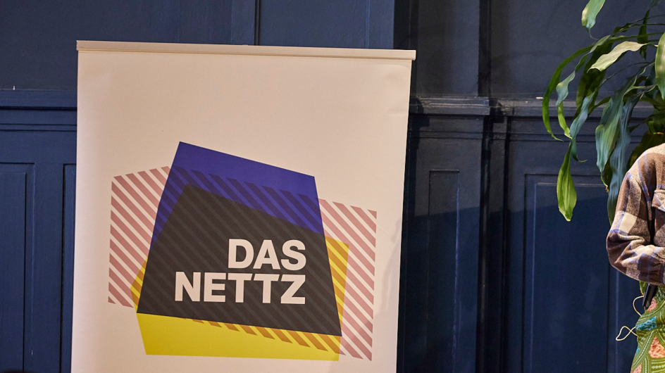 Es ist ein Roll-Up oder Banner zu sehen, auf dem Das NETTZ-Logo zu sehen ist. Es steht vor einer blauen Wand. 